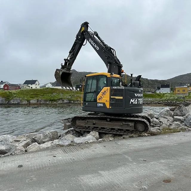 Gravemaskin fra MA TEK arbeider på steinfylling ved vannkant, med hus og åser i bakgrunnen.