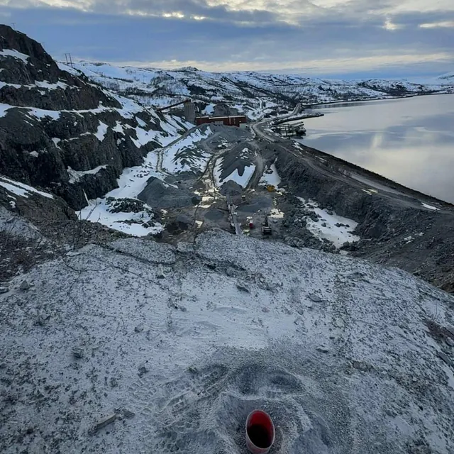 Utsikt over vinterlandskap med fjell og industriområde ved vann, rød plastrør stikker opp i forgrunnen.
