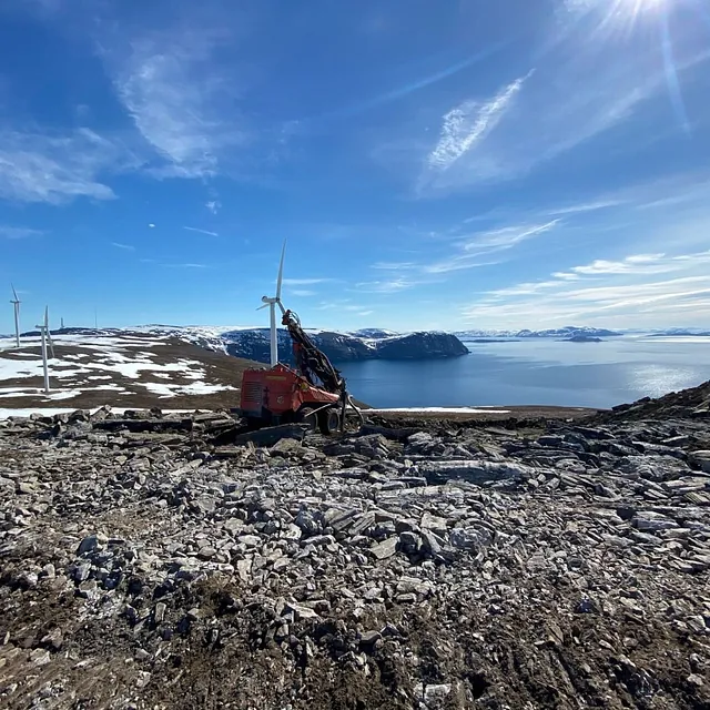 Gravemaskin på fjellterreng ved vindmøllepark, utsikt over sjø og snødekte fjell.