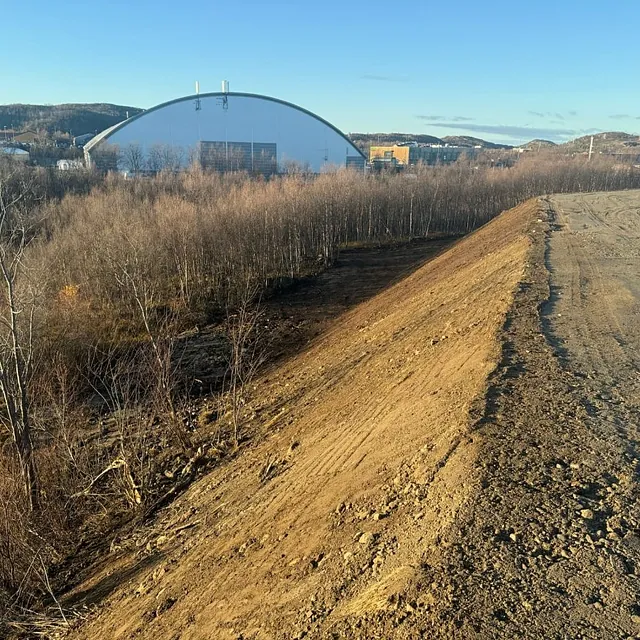 Utsikt fra en jordvoll ned mot skog og en stor buet hall i bakgrunnen.