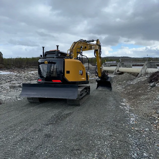 Gravemaskin fra CAT på gruslagt anleggsvei nær store betongrør i skogsområde.
