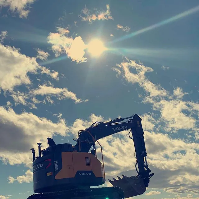 Gravemaskin står på steinhaug i motlys med sol og skyer i bakgrunnen.