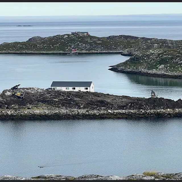 Anleggsmaskiner og hvitt bygg på liten øy omgitt av sjø og steinmolo.