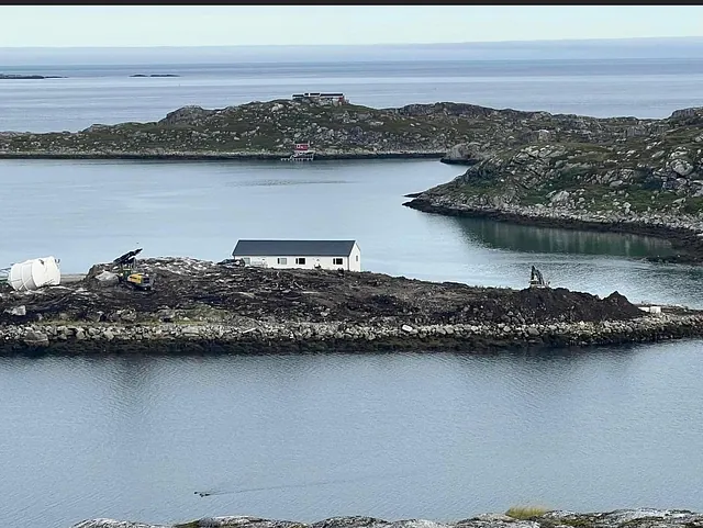 Anleggsmaskiner og hvitt bygg på liten øy omgitt av sjø og steinmolo.