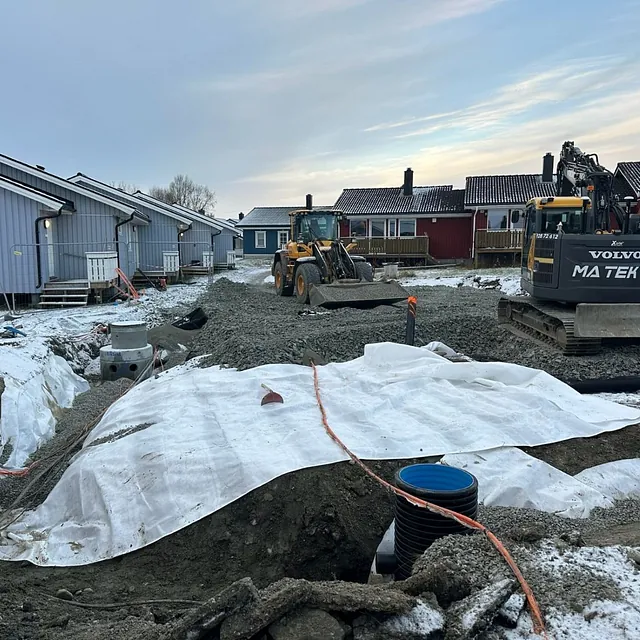 Anleggsområde med gravemaskiner og snødekt underlag ved rekkehus, arbeid pågår i grøft.