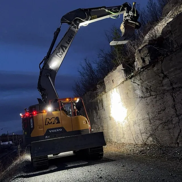 Gravemaskin lyser opp fjellvegg i skumringen under arbeid på grusvei.