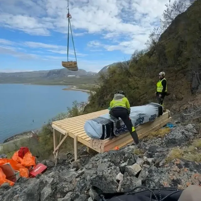Arbeidere på platting i bratt fjellside, helikopterløft frakter last med utsikt mot fjord.