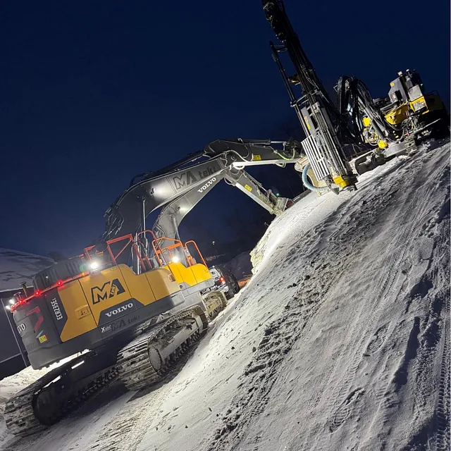 Gravemaskin og borerigg i arbeid i mørket, på snødekt skråning med spor i bakken.
