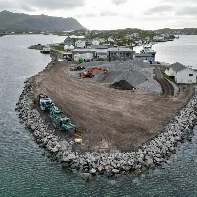Landfylling og anleggsarbeid på molo, med maskiner og grus i havneområde.