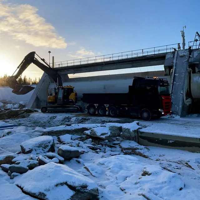 Gravemaskin og lastebil i snødekt landskap ved bru, med teksten «Melkefoss».