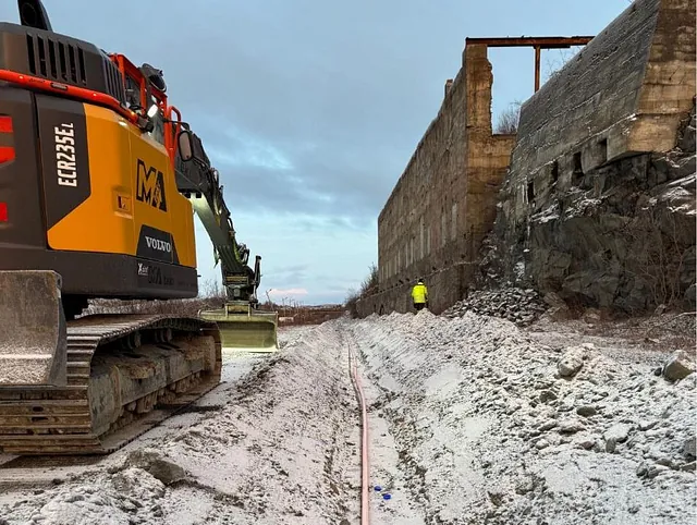 Grøft med rør og gravemaskin ved murbygg, arbeider med refleksjakke i bakgrunnen.