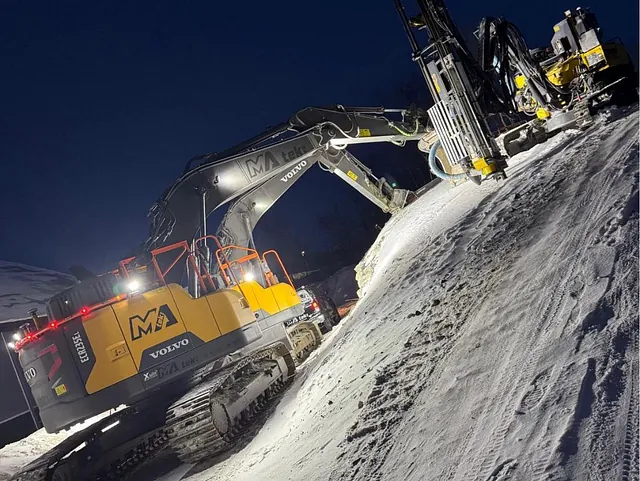 Gravemaskin og borerigg i arbeid i mørket, på snødekt skråning med spor i bakken.